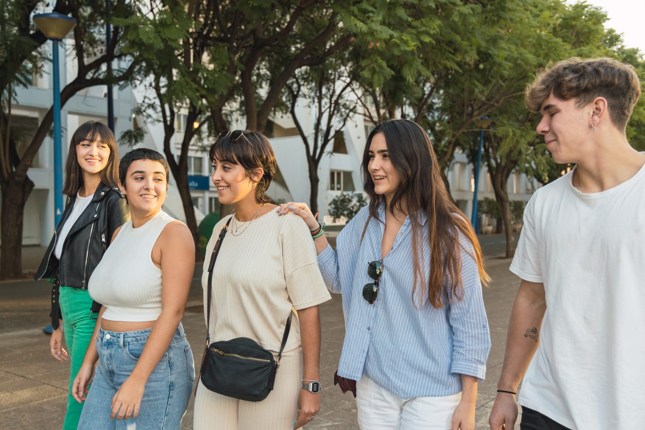 Group of diverse friends walking on street near green trees under blue sky